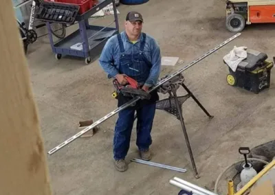 A man standing in a workshop looking at a long piece of metal material secured in a vice on a workbench, with a power tool in hand.