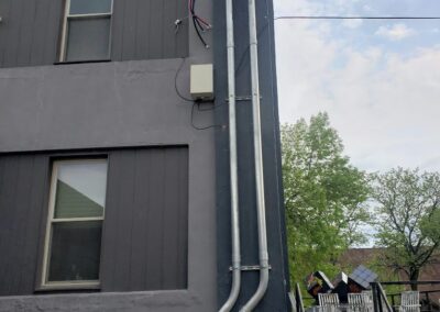 Gray building exterior with three windows and a cable conduit running along the side. Several cables extend from the top conduit. Trees and an outdoor seating area are visible in the background.