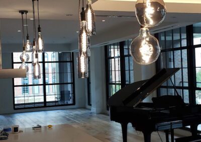 Modern kitchen and living area with large windows and hanging light fixtures. A black grand piano is placed near the windows. The space is minimalistic with white countertops.