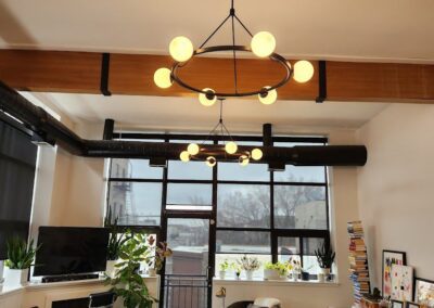 A well-lit living room with modern chandeliers, a couch, a TV, multiple plants, a tall stack of books, and a coffee table with various items on it. Large windows let in natural light.