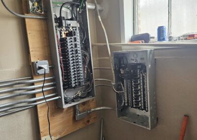 An electrical panel setup in a partially finished room with wires running to and from two circuit breaker boxes mounted on the wall near a small window.